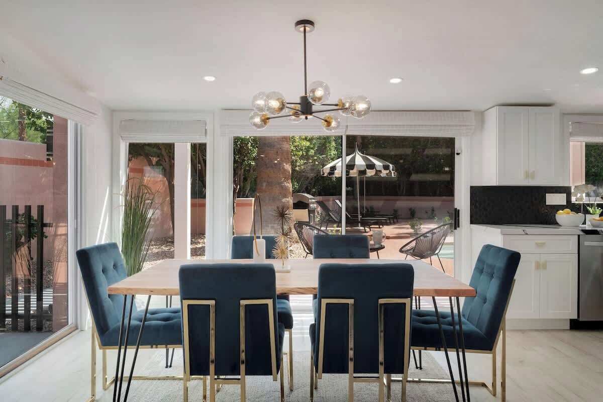 A dining area with a wooden table and blue upholstered chairs, a modern chandelier, and a kitchen area with white cabinets and a black countertop.