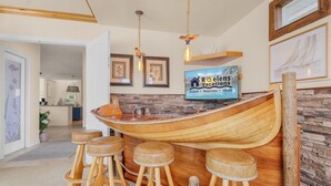 Un coin bar en bois avec quatre tabourets, un téléviseur diffusant des images promotionnelles de « Roelens Vacation », et des œuvres d’art encadrées accrochées aux murs.