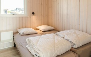 Un lit simple avec deux oreillers et une couverture, une table de chevet, une lampe fixée au mur et une fenêtre donnant sur les bâtiments environnants.