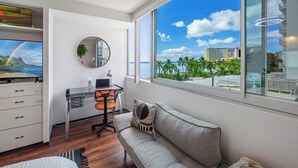 A modern hotel room with a large window offering a view of the beach and palm trees, a desk with a laptop, a comfortable sofa, and a TV displaying a scenic landscape. - Waikiki 1-bedroom condo with AC and wifi (Honolulu)