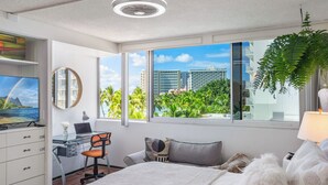 A modern hotel room with a large bed, a desk with a chair, a television, and a large window offering a view of palm trees and buildings. - Waikiki 1-bedroom condo with AC and wifi (Honolulu)
