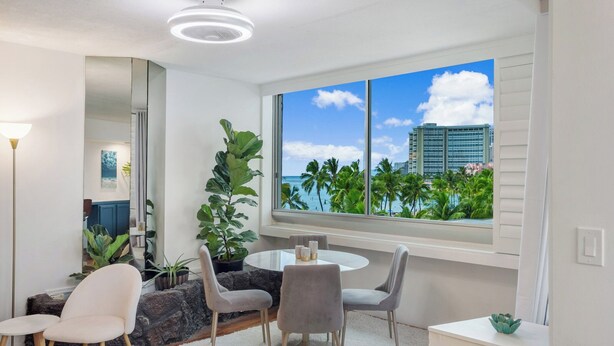 A modern dining area with a round table, four chairs, and a large window offering a view of palm trees and a city skyline. - Waikiki 1-bedroom condo with AC and wifi (Honolulu)