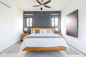 Une chambre moderne avec un lit en bois, deux tables de chevet avec des lampes, un ventilateur de plafond et un grand tableau abstrait accroché au mur.