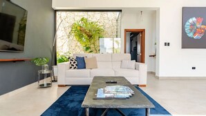 A modern living room with a white sofa, a coffee table with magazines, a large TV, and a stone wall with plants.