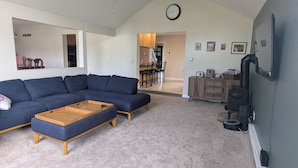 A living room with a sectional sofa, a wooden coffee table, and a kitchen area visible in the background.