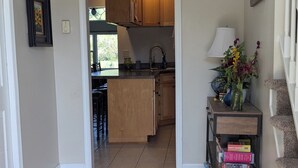 A kitchen with wooden cabinets, a sink, and a countertop. There is a vase with flowers on a shelf, and a cat is visible on the floor.