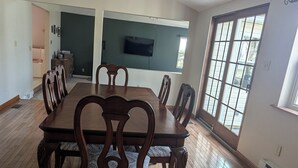 A spacious dining room with a large wooden table and chairs, a chandelier, and a television mounted on the wall.
