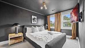 Un dormitorio con una cama grande, una mesita de noche de madera, una lámpara negra, un cuadro enmarcado en la pared y una ventana con cortinas.
