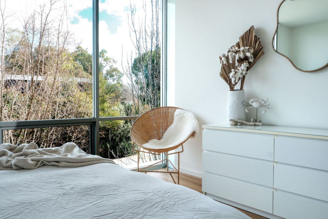 A bedroom with a large window, a bed, a white dresser, a wicker chair, and a decorative wall piece.