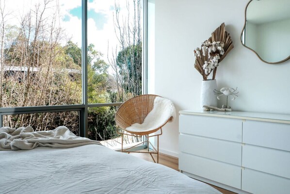 A bedroom with a large window, a bed, a white dresser, a wicker chair, and a decorative wall piece.