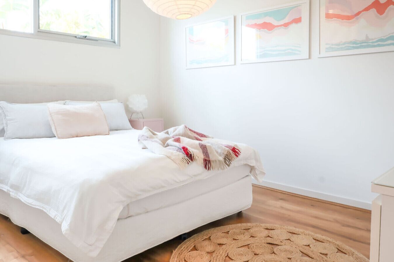 Una cama bien hecha con ropa de cama blanca y una colcha roja y blanca, una mesita de noche con una lámpara, y una alfombra tejida sobre el suelo de madera.