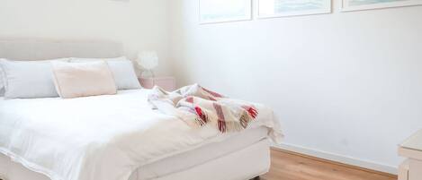Una cama bien hecha con ropa de cama blanca y una colcha roja y blanca, una mesita de noche con una lámpara, y una alfombra tejida sobre el suelo de madera.