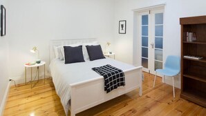 A bedroom with a bed, bedside tables, a chair, a bookshelf, and a door with frosted glass panels. - North Adelaide - Row Cottage - “Monte” (North Adelaide)