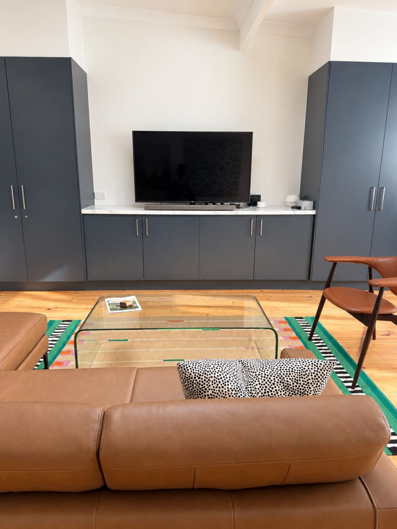 A modern living room with a leather sofa, a glass coffee table, a television on a stand, and grey cabinets.
