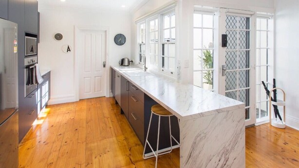 A modern kitchen with a marble island, wooden flooring, and a view through a glass door. - North Adelaide - Row Cottage - “Monte” (North Adelaide)