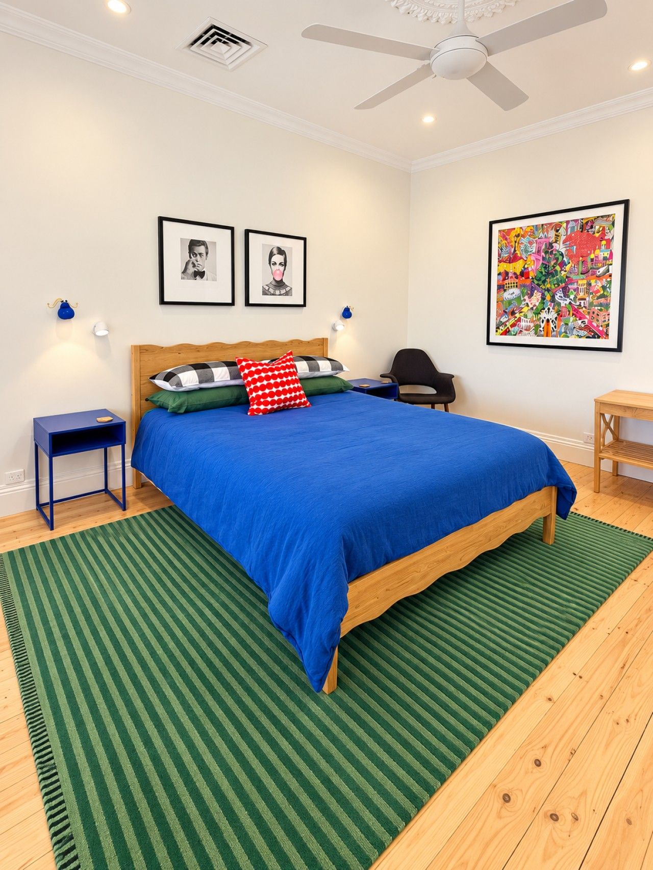 A bedroom with a wooden bed, green striped rug, two wall-mounted lamps, a ceiling fan, and two framed pictures.