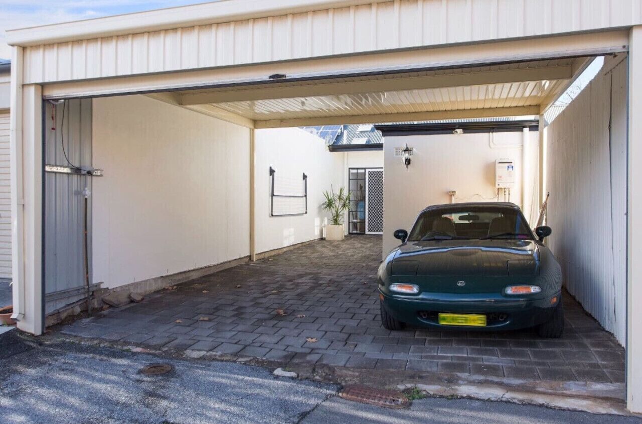 A car parked inside a garage with a white exterior, a brick floor, and a small plant in the corner.