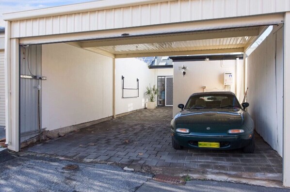 Ein Auto steht in einer Garage mit weißer Außenfarbe, einem Backsteinboden und einer kleinen Pflanze in der Ecke.