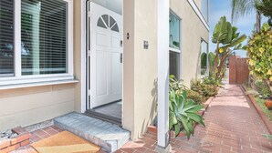 Uma entrada residencial com porta branca, um pequeno alpendre e um caminho de tijolos que leva a uma casa.
