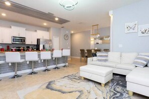 Una cocina moderna con armarios blancos, un microondas y un refrigerador. Hay cuatro taburetes blancos y una mesa de comedor con sillas. La sala de estar tiene un sofá blanco y una alfombra estampada.