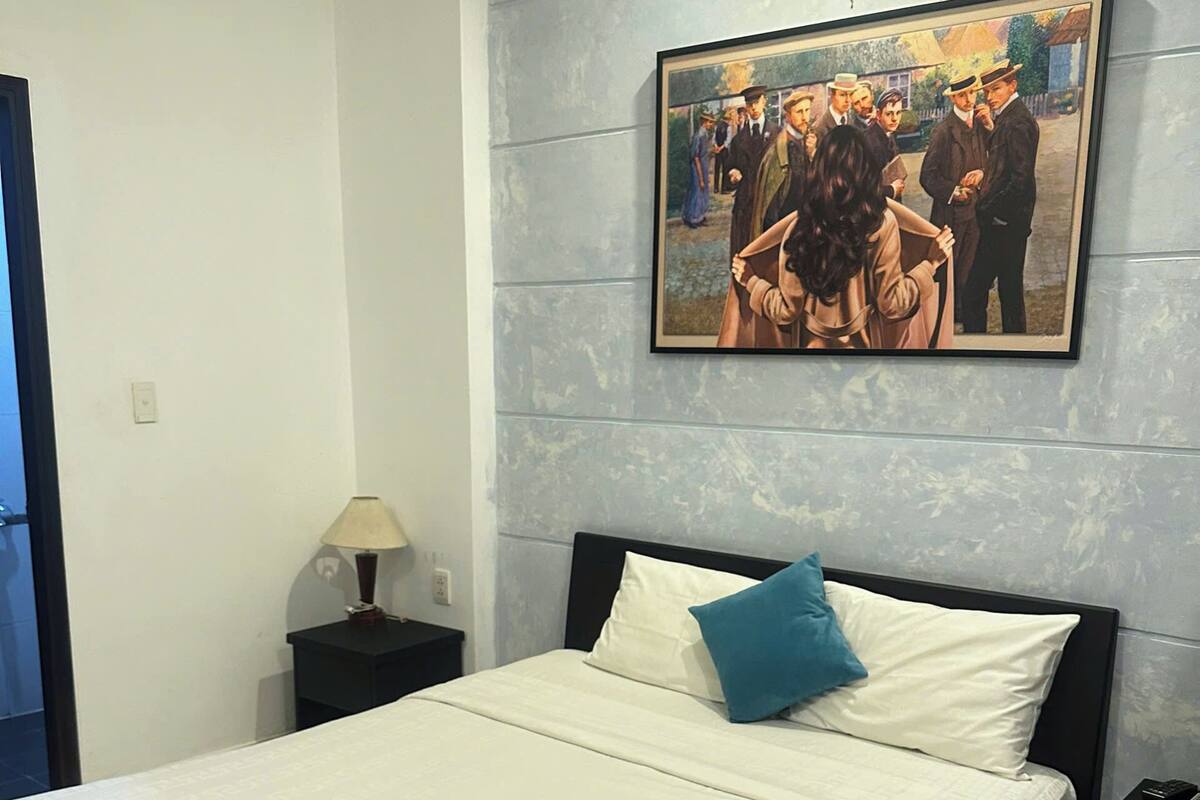 Ein ordentlich gemachtes Bett mit weißer Bettwäsche und einem blauen Kissen, ein gerahmtes Bild an der Wand und ein kleiner Nachttisch mit einer Lampe.