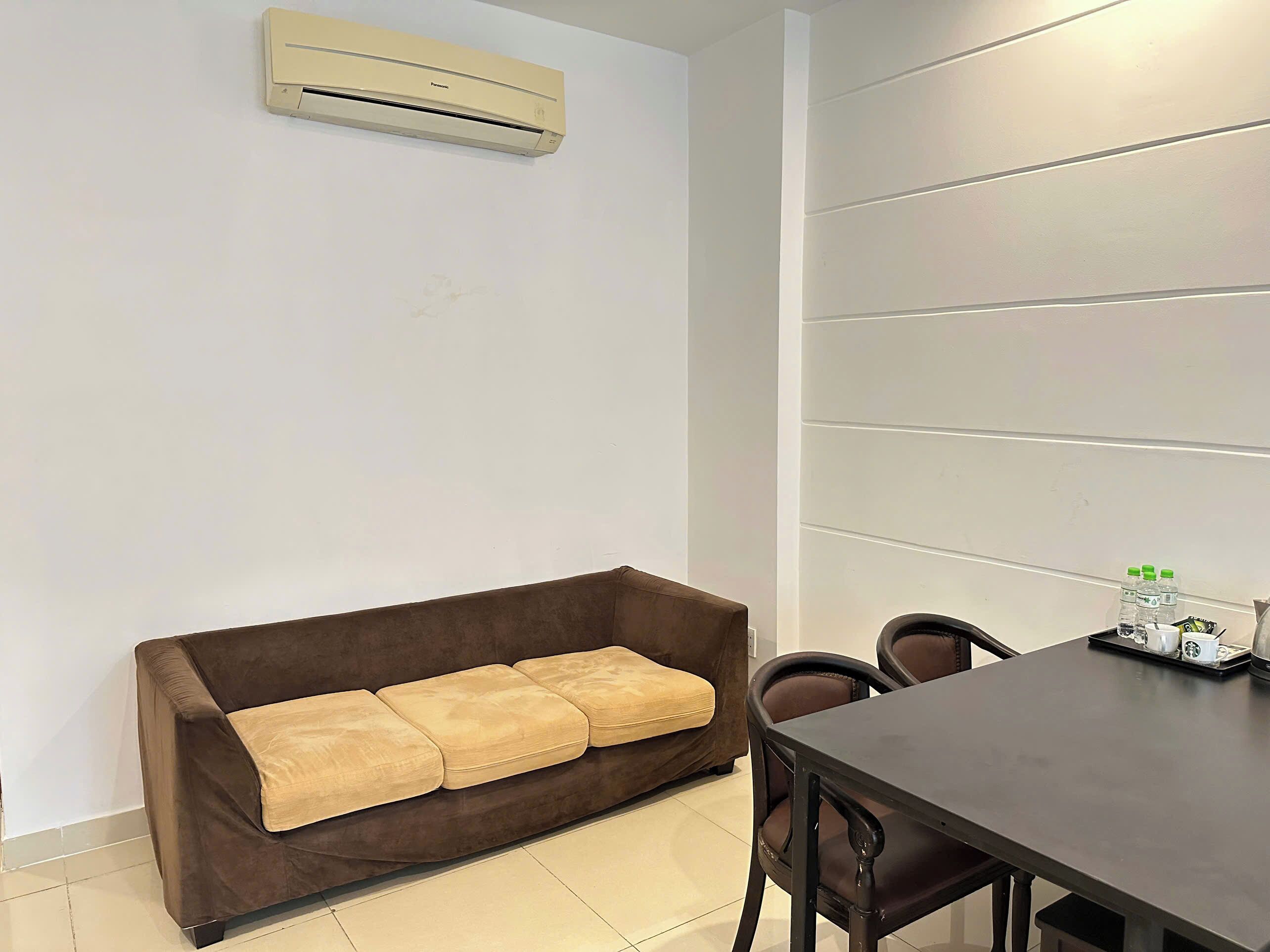 Une pièce avec un canapé marron, une table à manger avec des chaises, un climatiseur et un mur recouvert de panneaux horizontaux.