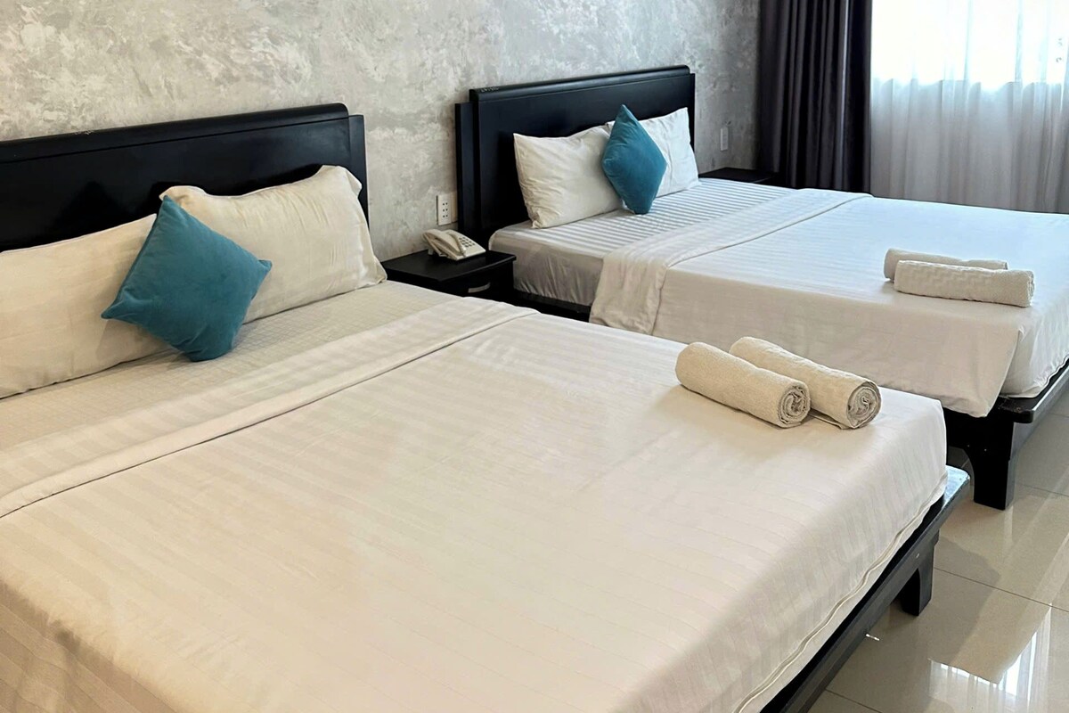 A hotel room with two beds, a concrete accent wall, a nightstand with a phone, and a window with curtains.