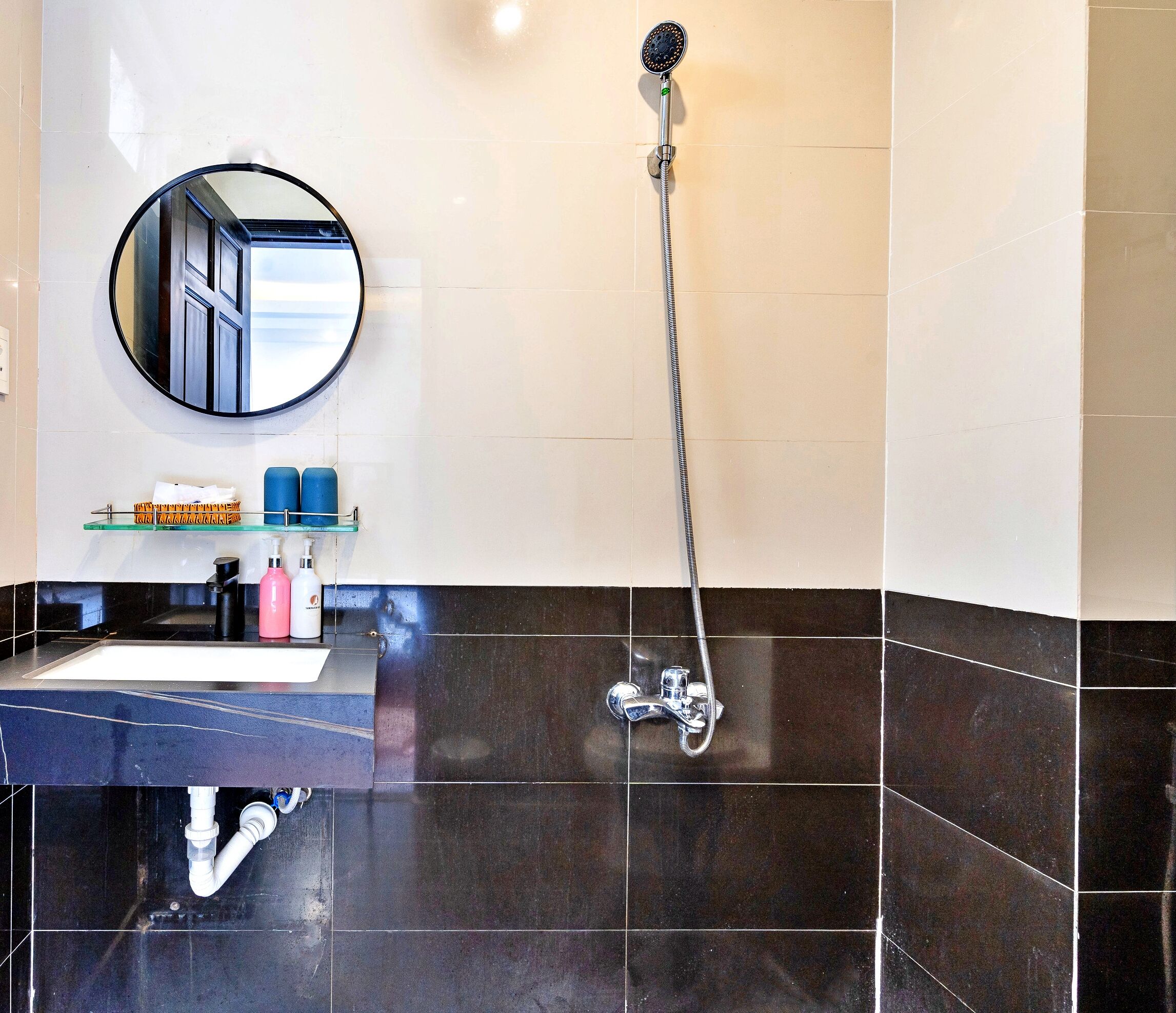Un baño con suelo de baldosas blanco y negro, un espejo redondo, un lavabo con repisa de vidrio encima y una ducha con cabezal de mano.