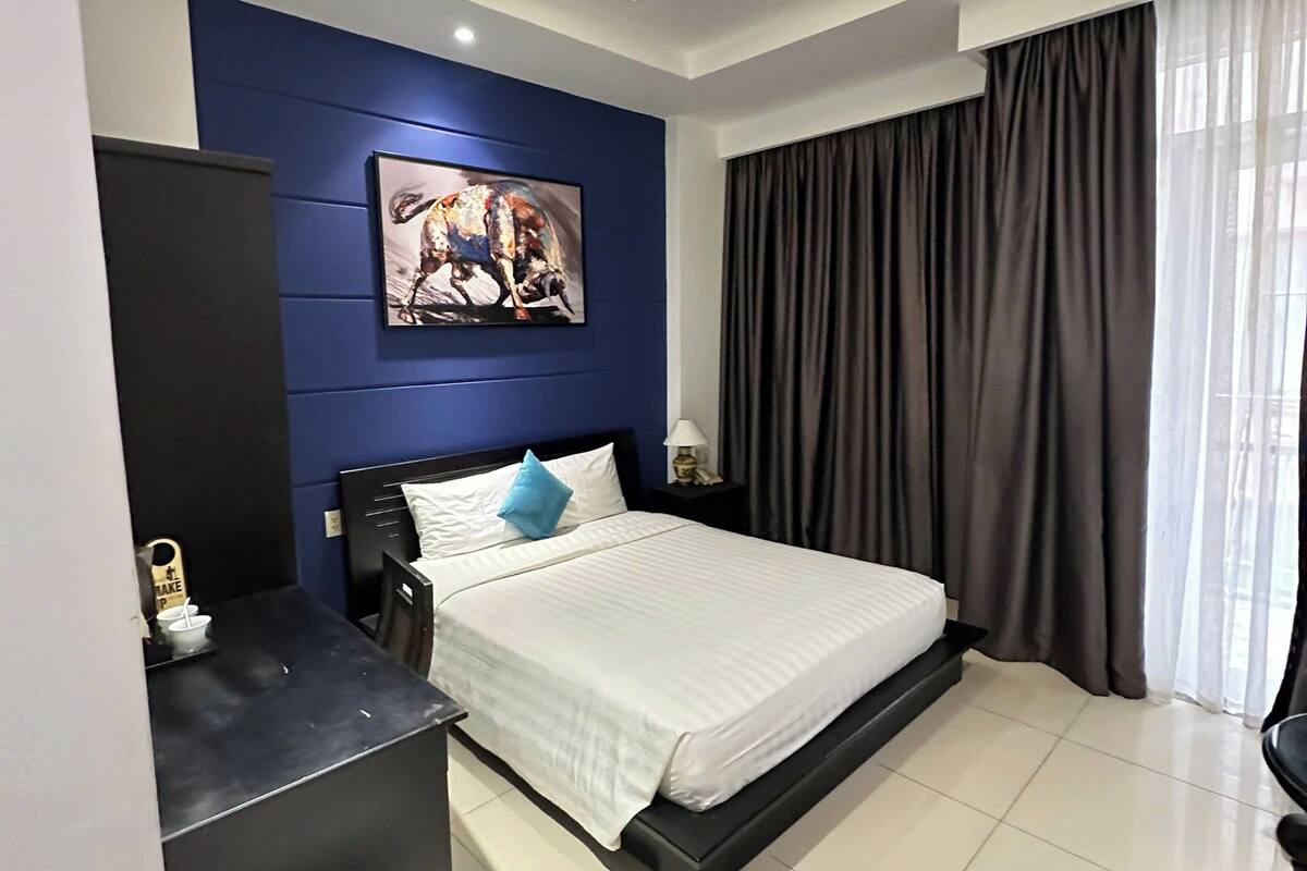 Ein modernes Hotelzimmer mit Bett, Schreibtisch, Stuhl, kleinem Kühlschrank, Nachttisch mit Lampe, einem Bild an der Wand und einem großen Fenster mit Vorhängen.