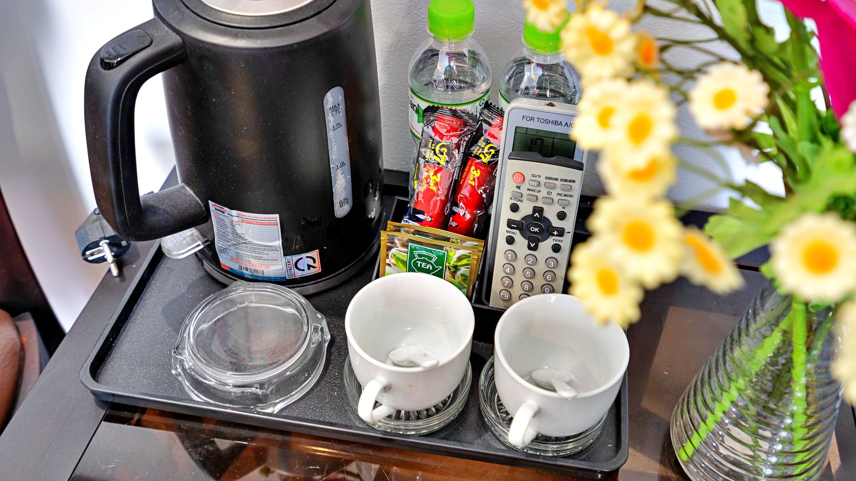 Una cafetera, dos tazas, una bolsita de té, una botella de agua y un control remoto sobre una bandeja.