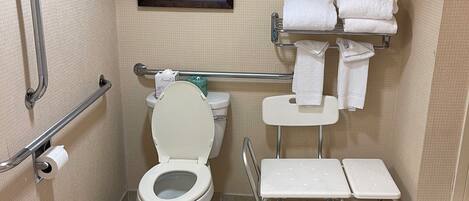 A bathroom with a toilet, handrails, a sink, a mirror, and a shelf with towels.