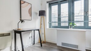 Een kamer met een bureau, een staande lamp, een radiator en een raam met uitzicht op gebouwen.