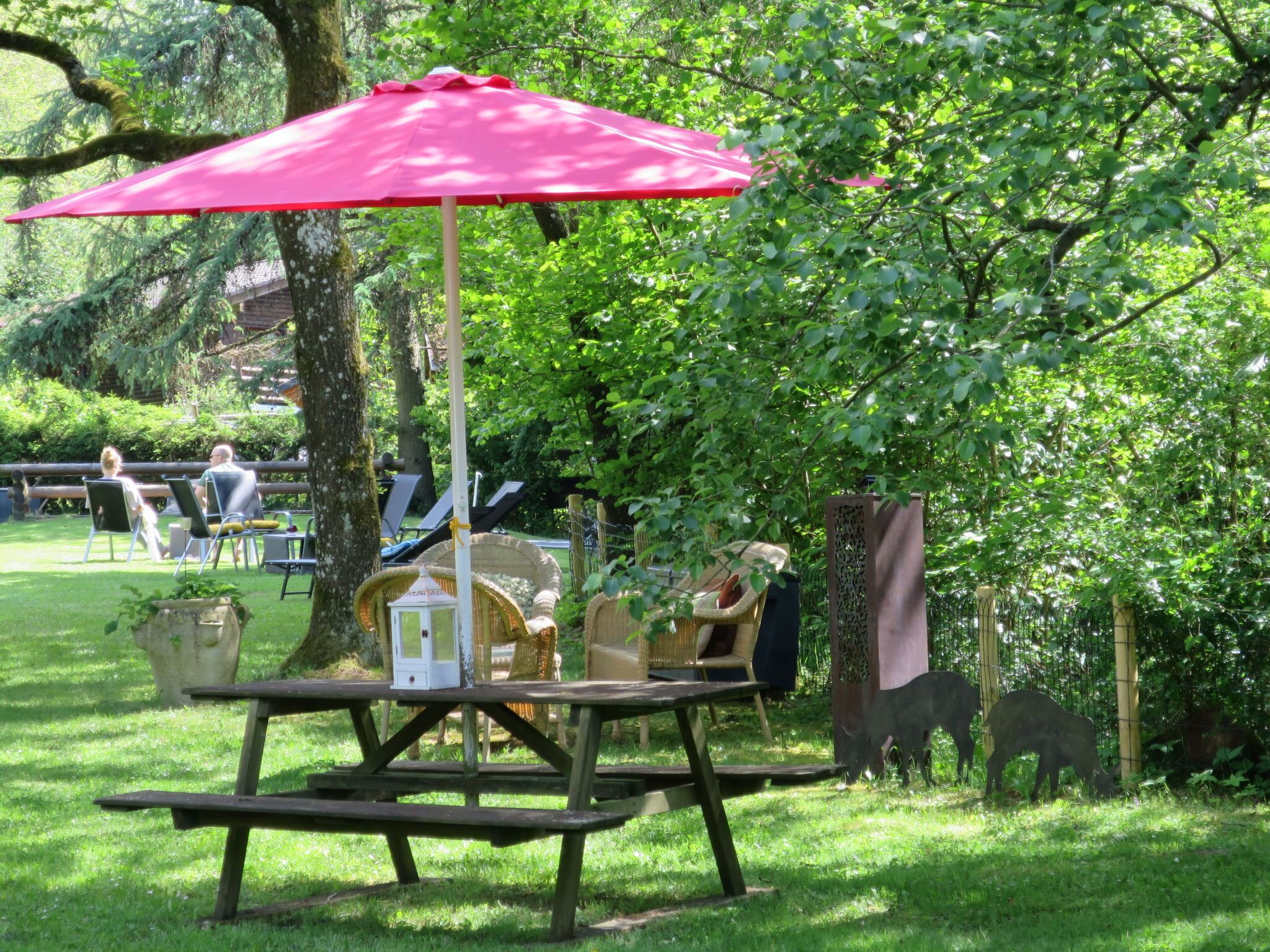Ein Picknicktisch mit rotem Sonnenschirm, umgeben von Grünpflanzen und einem Zaun.
