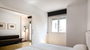 Une chambre à coucher avec un grand lit, un canapé sombre, une lampe sur pied et une fenêtre donnant sur des immeubles.