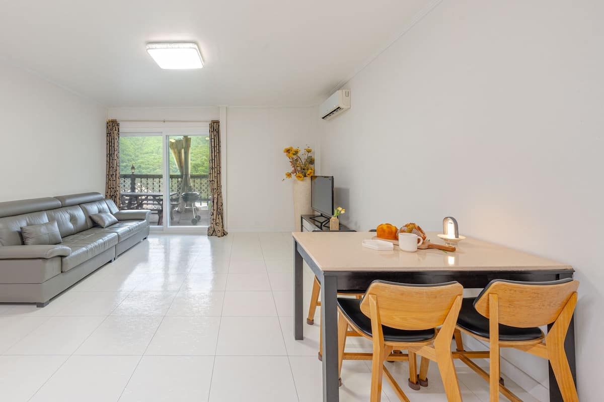 A modern living room with a grey sofa, a dining area with a table and chairs, and a view of a balcony.