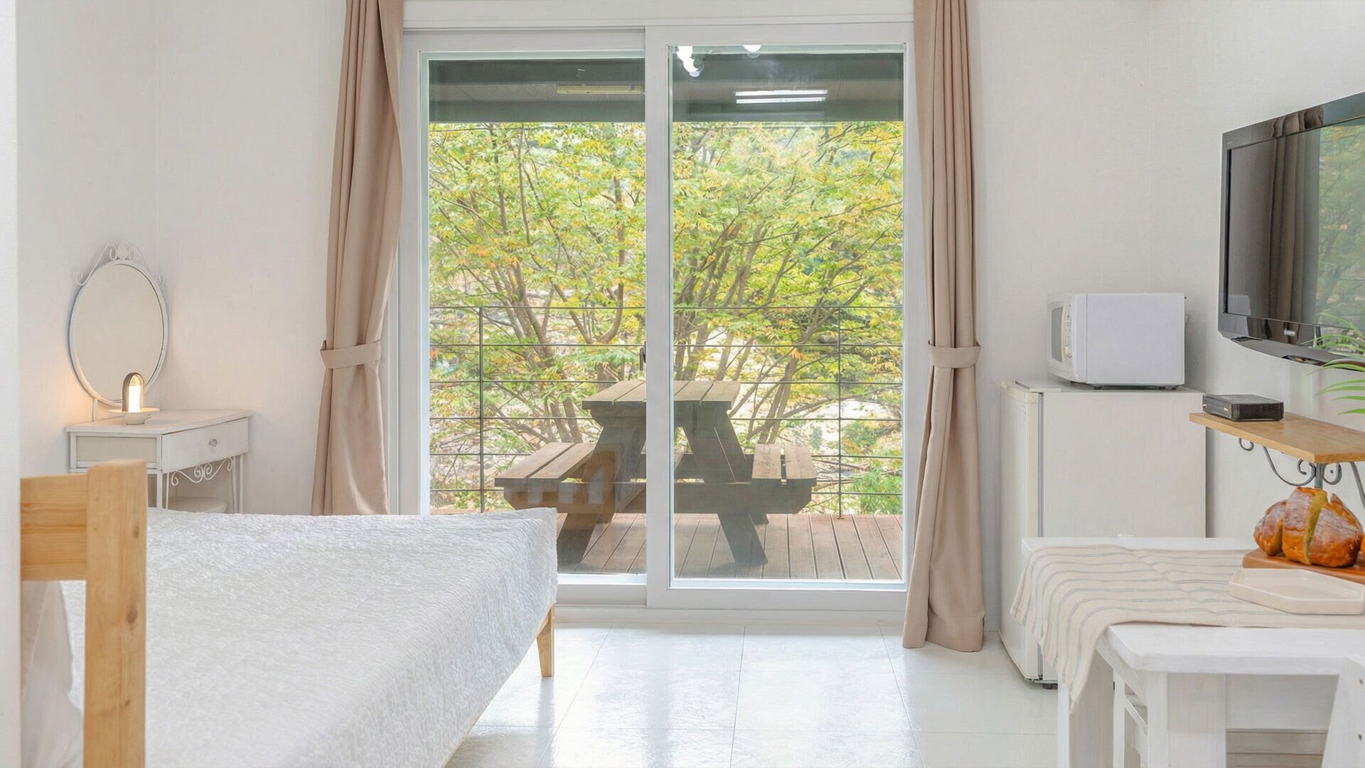 Ein Schlafzimmer mit Bett, Nachttisch mit Lampe, Fernseher und Blick auf einen Balkon mit Tisch und Stühlen.