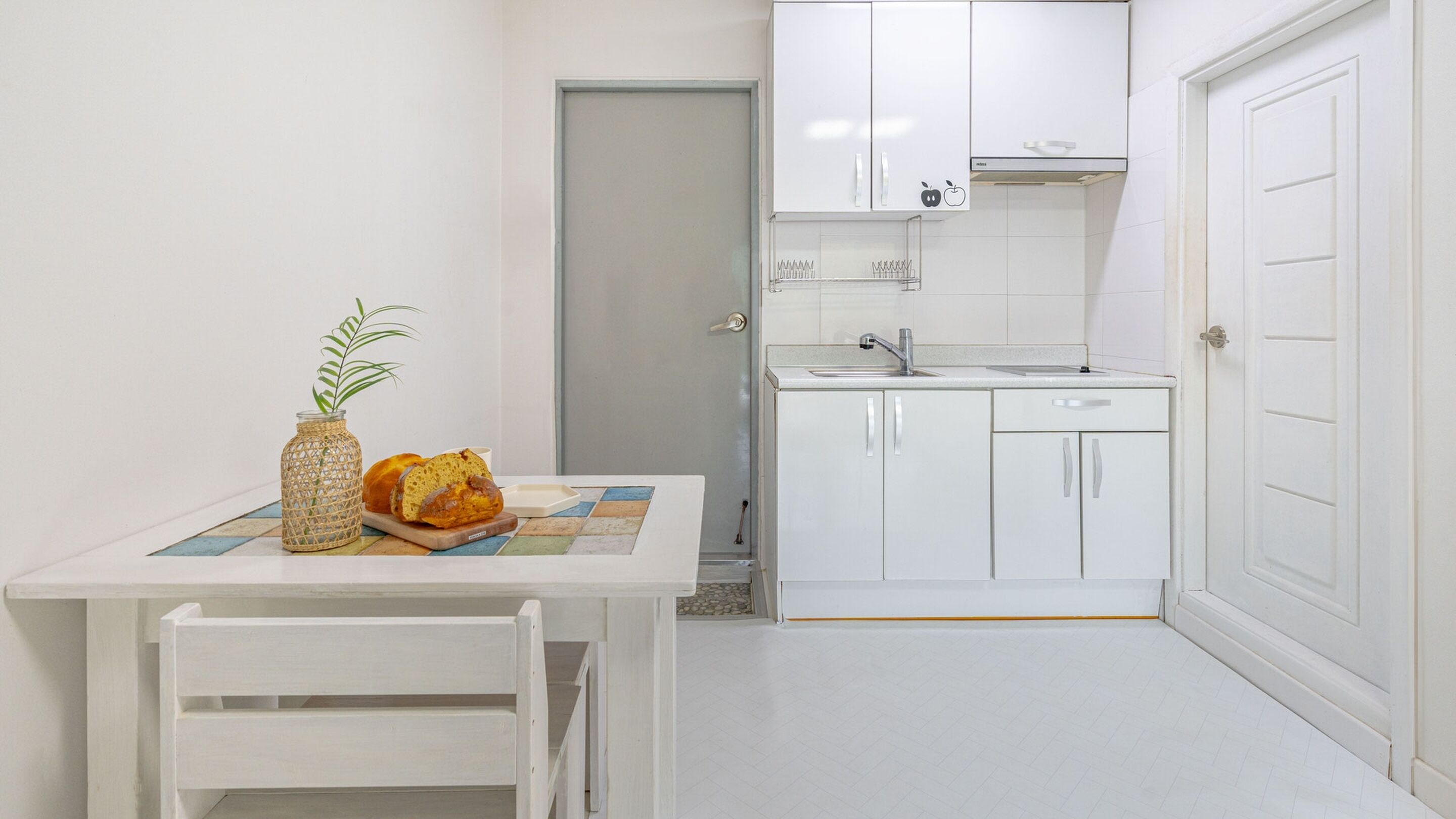 A small, white kitchen with a single sink, a white door, and a tiled backsplash.
