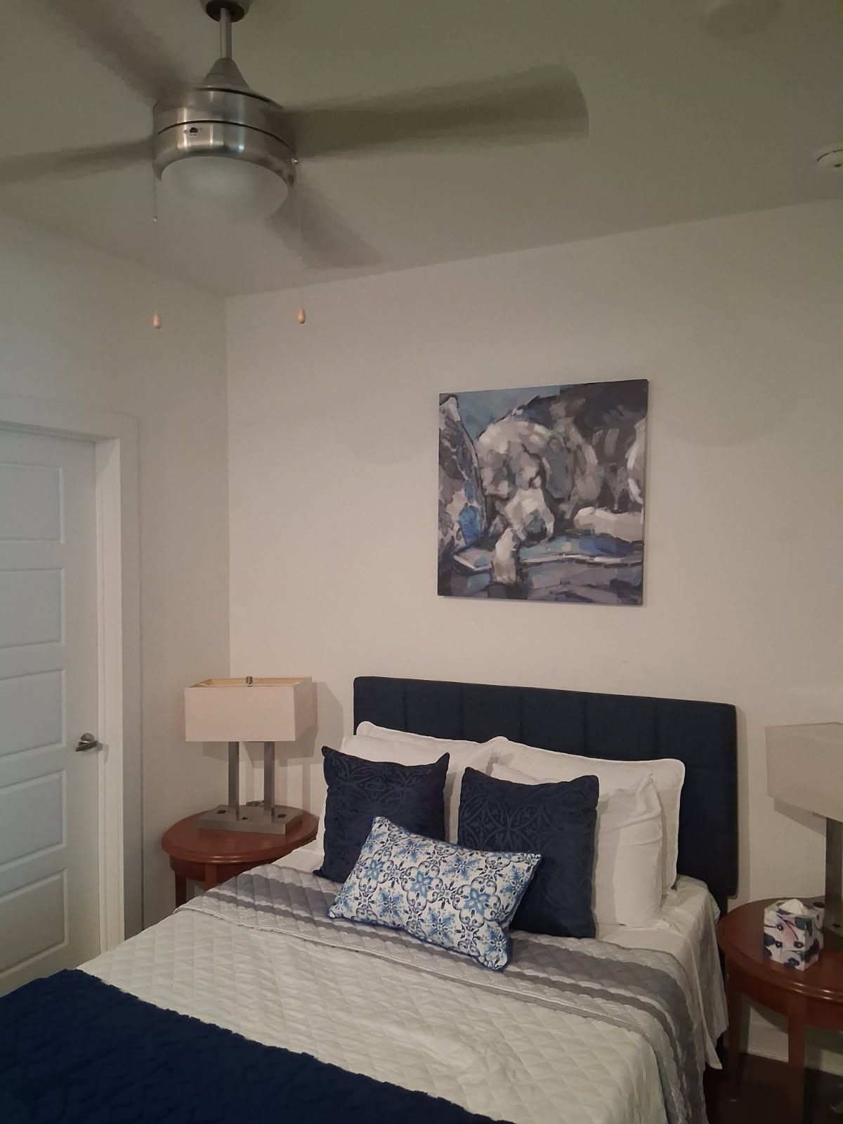 Een slaapkamer met een bed, een plafondventilator, een schilderij en een nachtkastje met een lamp.