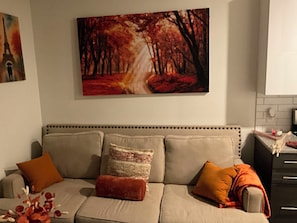 A living room with a sofa, cushions, a painting, and a kitchen area.