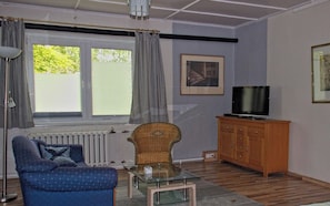 Sala de estar com sofá, poltrona, mesa de centro de vidro, aparador de madeira, televisão e janela com cortinas.