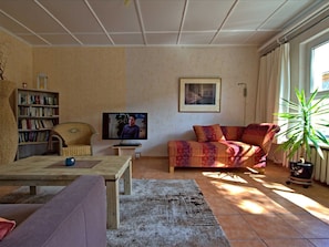 Uma sala de estar com sofá, mesa de centro, estante de livros, televisão e uma planta em vaso.