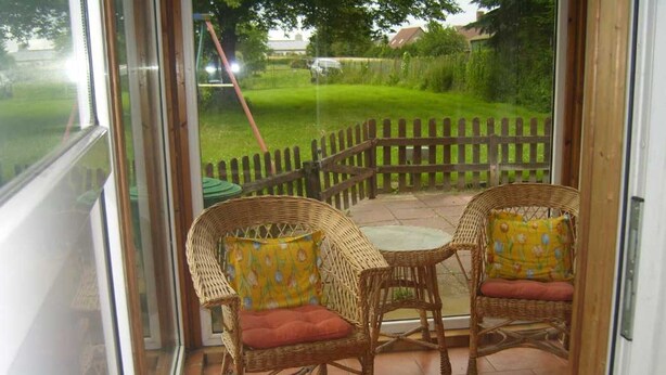 A patio with wicker chairs and a table, a glass door leading outside, and a view of a grassy yard with a wooden fence. - Apartment 'Kelpin' with Private Terrace and Wi-Fi (Möllenbeck)