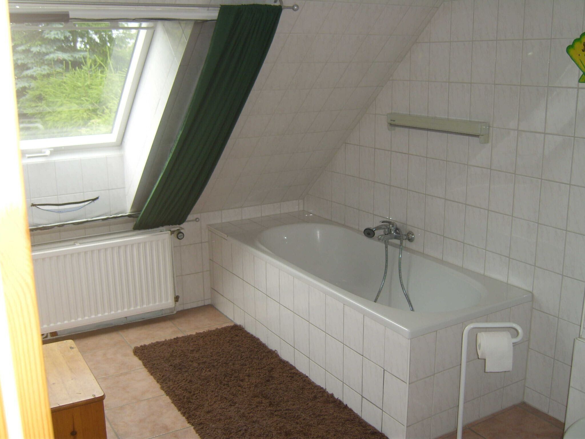A small, clean bathroom with a bathtub, a window with a green curtain, white tiled walls, and a brown rug.