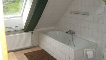 A small, clean bathroom with a bathtub, a window with a green curtain, white tiled walls, and a brown rug.