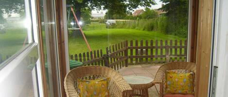 A glass-enclosed patio with wicker chairs and cushions, a tiled floor, and a view of a green lawn and wooden fence.
