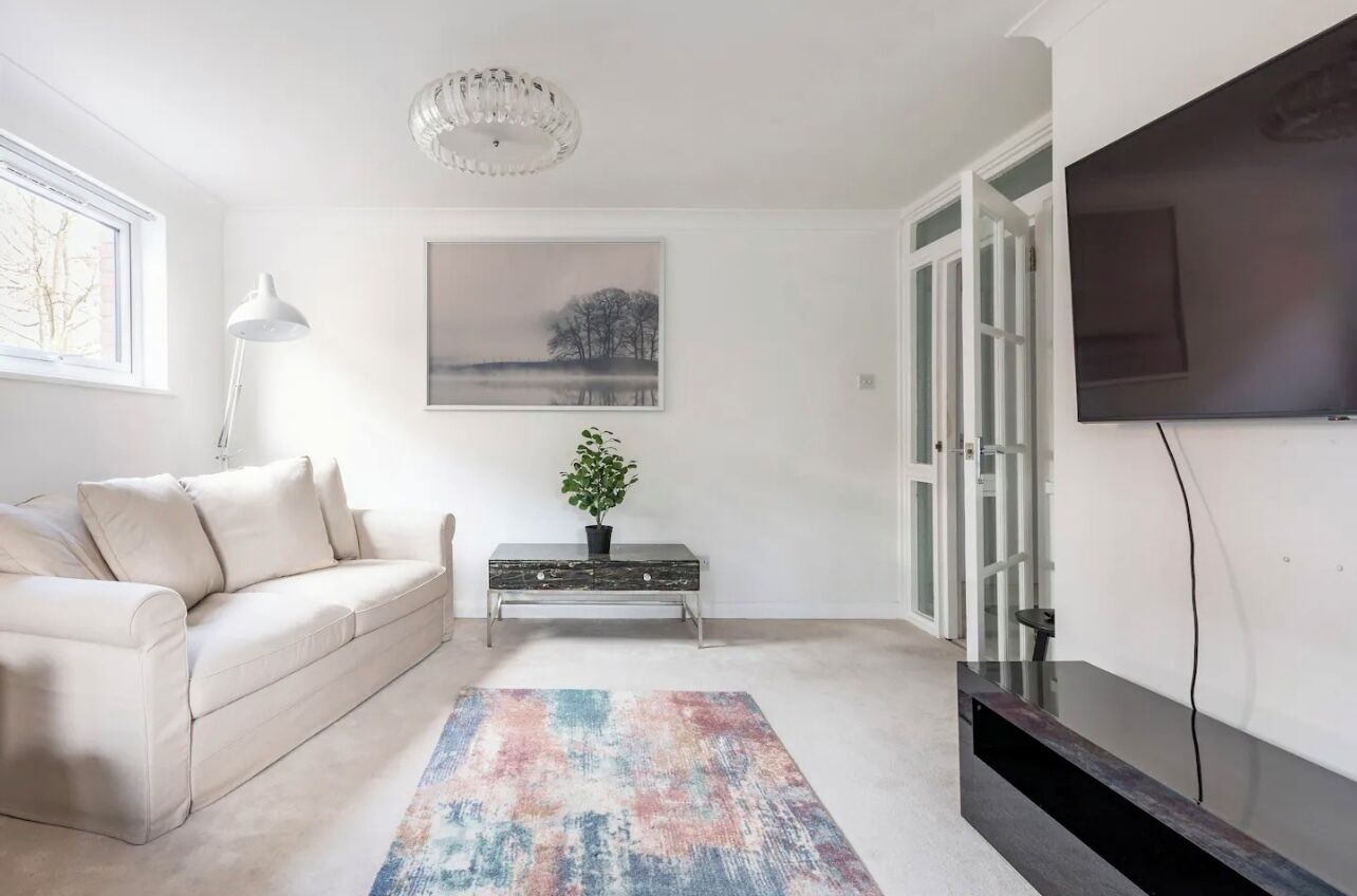 Un salon avec un canapé beige, un tapis aux couleurs vives, un tableau représentant un paysage, une plante en pot et un téléviseur à écran plat.