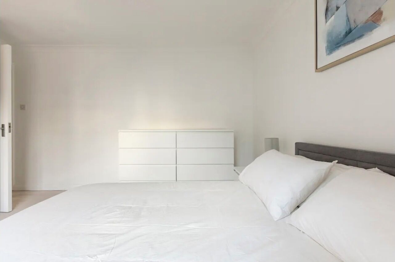 Ein schlicht eingerichtetes Schlafzimmer mit einem großen Bett, weißer Bettwäsche, einem grauen Kopfteil und einem Nachttisch in Weiß mit einer Lampe.