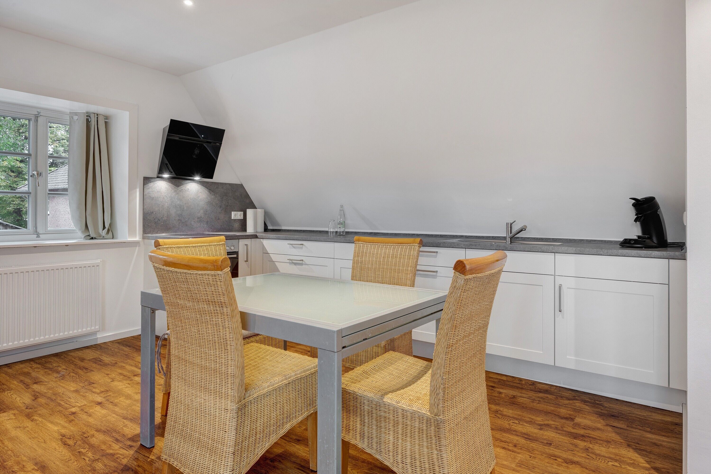 A dining area with a table and chairs, a kitchen in the background, and a window with curtains.