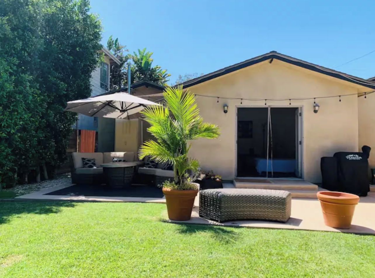 Un jardin à l’arrière d’une maison, aménagé avec une terrasse, du mobilier d’extérieur, un pot de fleurs et une porte-fenêtre coulissante donnant sur un espace abrité.