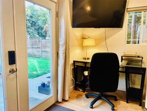 Ett rum med en skjutdörr i glas som leder ut till en trädgård, en svart kontorsstol, ett skrivbord med en lampa och en platt-tv som är monterad på väggen.